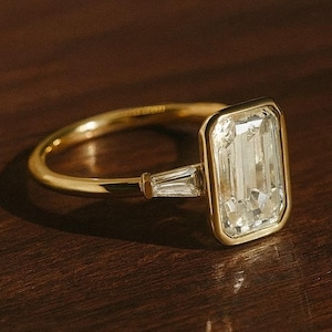 May include: A gold ring with a large, rectangular, clear gemstone as the focal point. The ring has a simple band and a smaller, rectangular gemstone on either side of the main stone. The ring is set against a dark brown background.