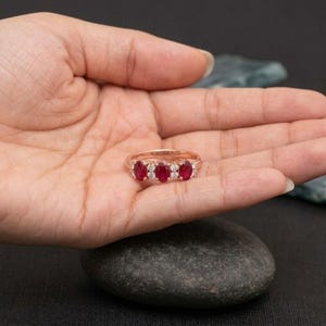 May include: A rose gold ring featuring three oval-cut red rubies arranged horizontally, separated and flanked by small, round, clear white diamonds set along the band. The metal has a warm, pinkish metallic luster throughout the design.