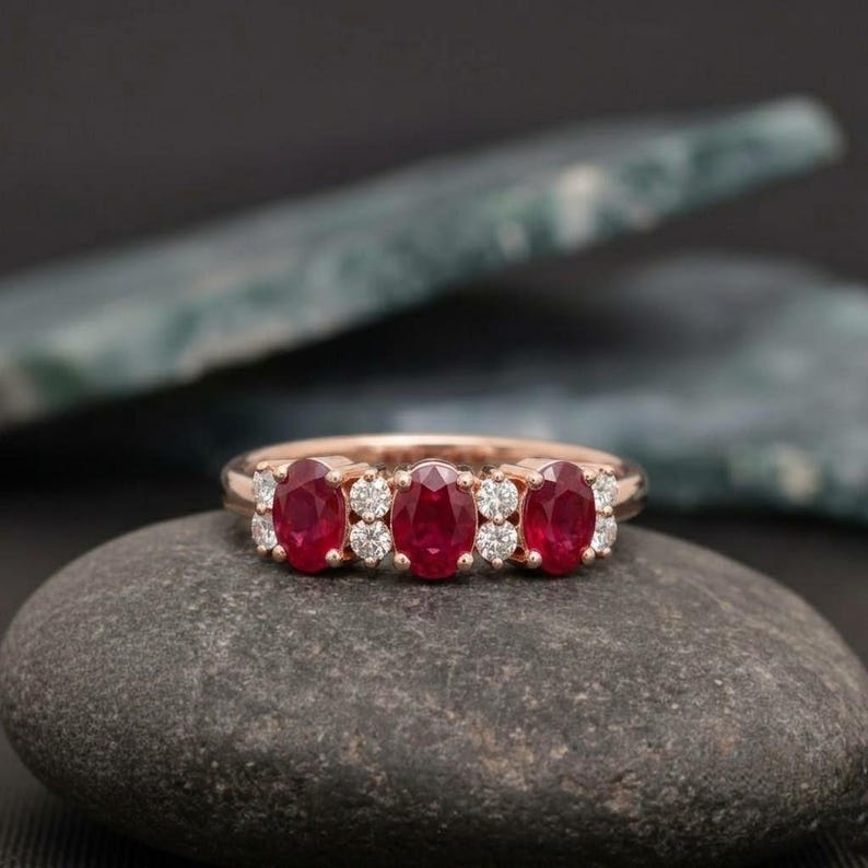 May include: A rose gold ring featuring three oval-cut red rubies arranged horizontally, interspersed with four small round-cut white diamonds. The band is polished metal, showcasing a classic multi-stone design with a vibrant color contrast.