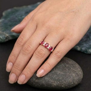May include: A rose gold ring featuring three oval-cut red rubies alternating with two small round-cut white diamonds. The gemstones are set in a horizontal row along the band, creating a classic multi-stone design with a polished metal finish.