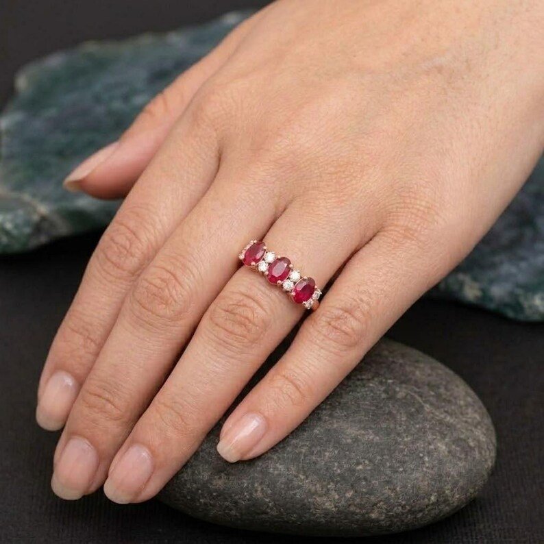 May include: A rose gold ring featuring three oval-cut red rubies alternating with two small round-cut white diamonds. The gemstones are set in a horizontal row along the band, creating a classic multi-stone design with a polished metal finish.