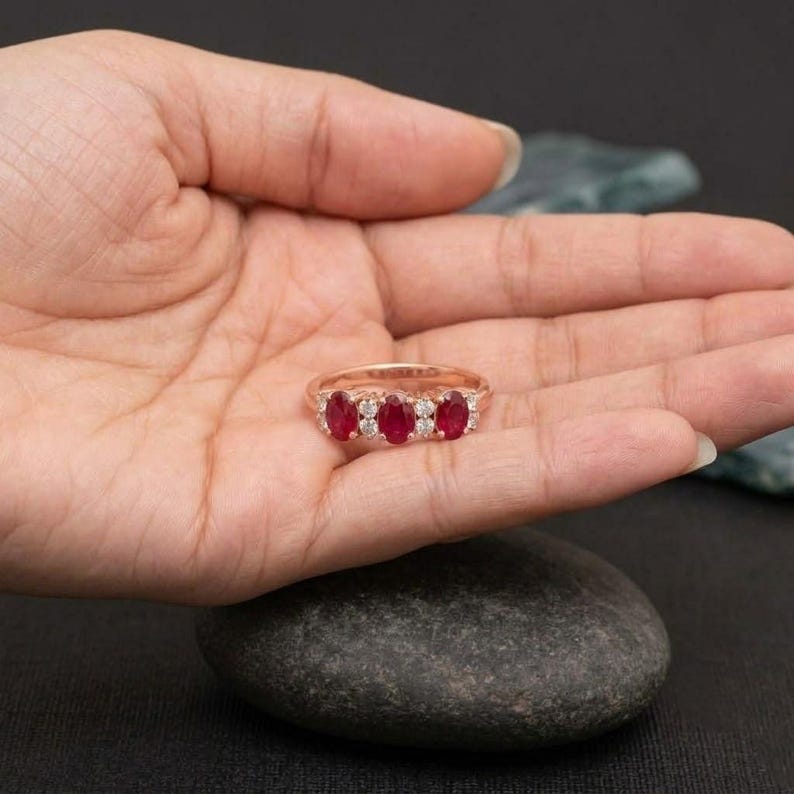 May include: A rose gold ring featuring three oval-cut red rubies arranged horizontally, separated and flanked by small, round, clear white diamonds set along the band. The metal has a warm, pinkish metallic luster throughout the design.