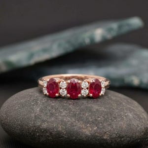 May include: A rose gold ring featuring three oval-cut red rubies arranged horizontally, interspersed with four small round-cut white diamonds. The band is polished metal, showcasing a classic multi-stone design with a vibrant color contrast.