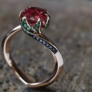 May include: A rose gold ring featuring a large, round, faceted red gemstone held by prongs shaped like leaves. The setting is accented with small, round green stones along the leaf-like prongs and a row of tiny black stones along the band.