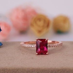 Puede incluir: Un anillo de oro rosa con una gema rectangular de color rojo intenso. La banda está adornada con pequeñas piedras claras. El anillo se encuentra sobre un fondo neutro con elementos florales borrosos.