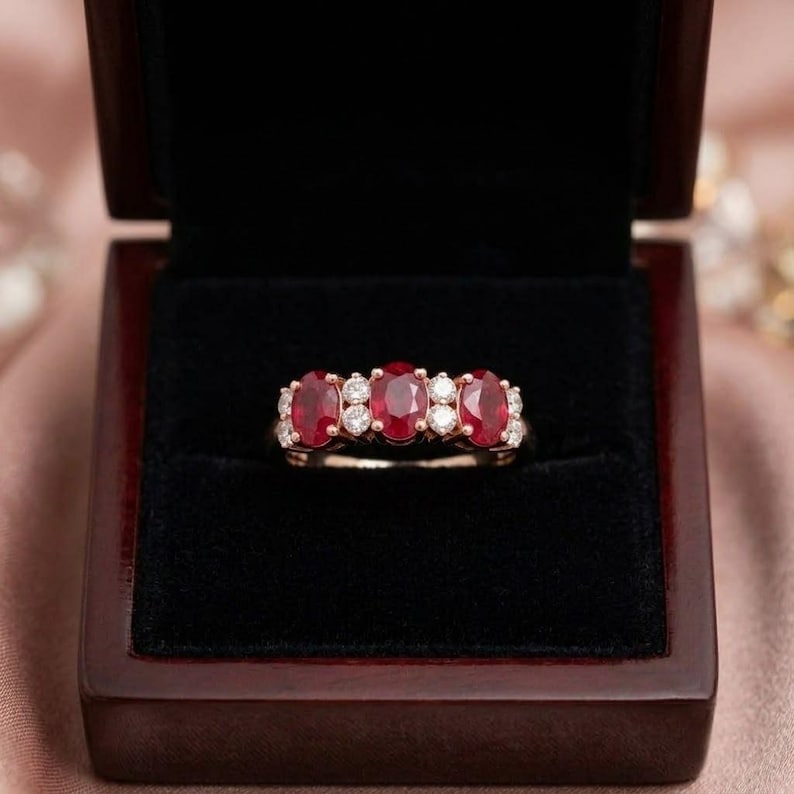 May include: A ring featuring three oval-cut red rubies and four round-cut white diamonds set in a polished metal band. The jewelry piece is displayed centered within a dark, velvet-lined wooden box with a hinged lid.