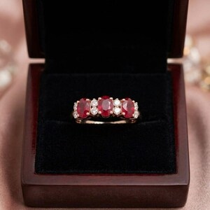 May include: A ring featuring three oval-cut red rubies and four round-cut white diamonds set in a polished metal band. The jewelry piece is displayed centered within a dark, velvet-lined wooden box with a hinged lid.
