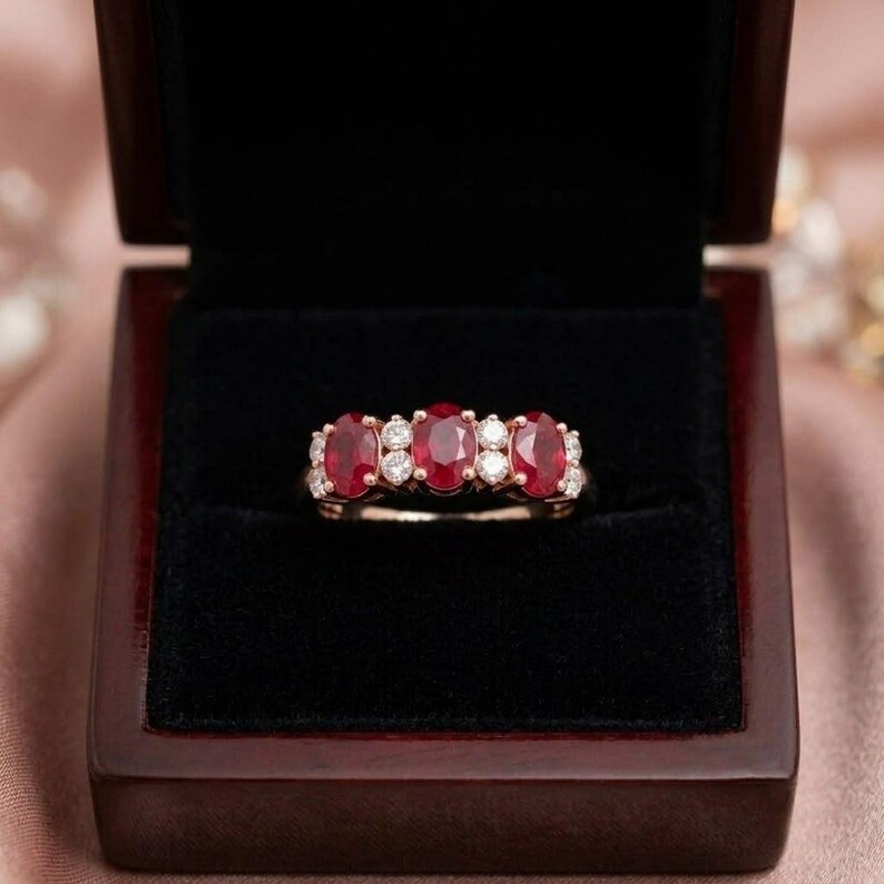 May include: A ring featuring three oval-cut red rubies and four round-cut white diamonds set in a polished metal band. The jewelry piece is displayed centered within a dark, velvet-lined wooden box with a hinged lid.