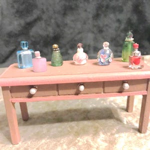 Pode incluir: Uma coleção de frascos de perfume em miniatura de várias formas e cores, expostos numa pequena mesa de toucador de madeira rosa com três gavetas. Os frascos incluem desenhos azuis, rosa, verdes e florais, com tampas decorativas.