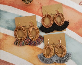 May include: Three pairs of oval-shaped earrings with woven wood and tassel accents. The tassels are black, rainbow, and grey. The earrings are labelled "knotsincards by nicole".