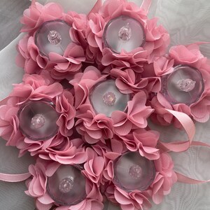 May include: Six pink floral LED lights, each with a clear plastic centre and layered fabric petals. Each light has a pink ribbon loop. The lights are designed for decorative purposes. The lights are approximately 10 cm in diameter.