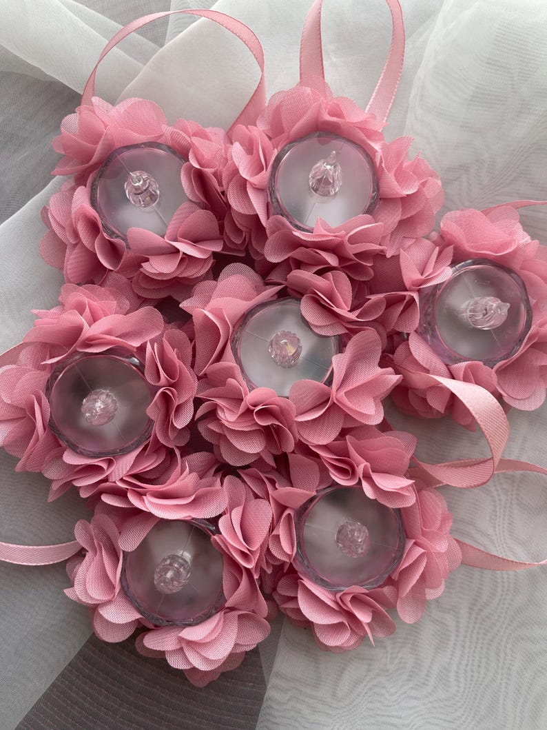 May include: Six pink floral LED lights, each with a clear plastic centre and layered fabric petals. Each light has a pink ribbon loop. The lights are designed for decorative purposes. The lights are approximately 10 cm in diameter.