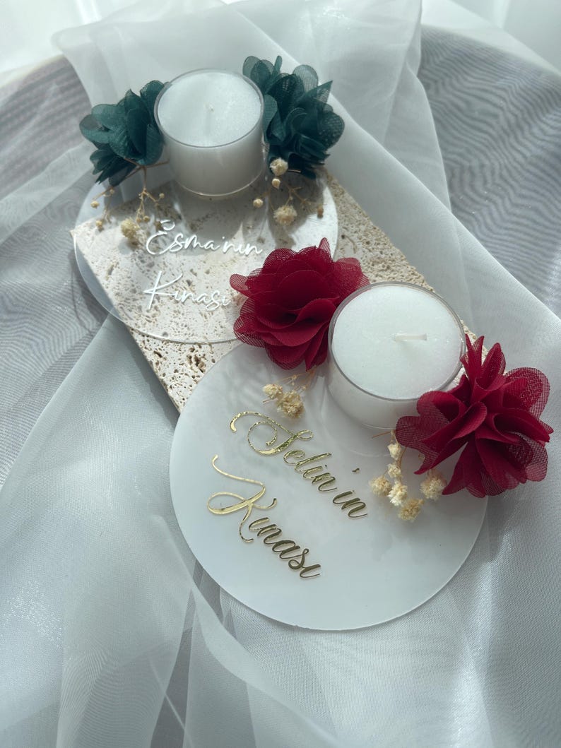 Op de afbeelding: Twee heldere acryl kaarshouders met witte theelichtjes, versierd met rode en groene bloemen van stof en gedroogde bloemen. De houders hebben de tekst "Esma'in Kinasi" en "Selin'in Kinasi" erop geschreven.