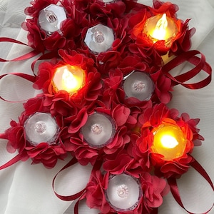May include: A collection of red fabric flower decorations, each with a clear, faceted, candle-shaped light. Some lights are illuminated with a warm, yellow glow, while others are unlit. Red ribbon loops are attached to the flowers.