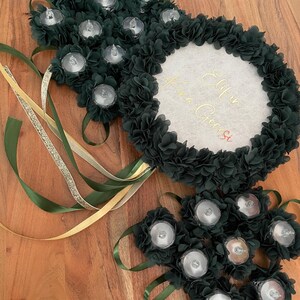 May include: Dark green floral arrangement with battery-operated lights and ribbons. The arrangement includes a central circular element with gold script. The design incorporates multiple clusters of dark green fabric flowers, each holding a small light.