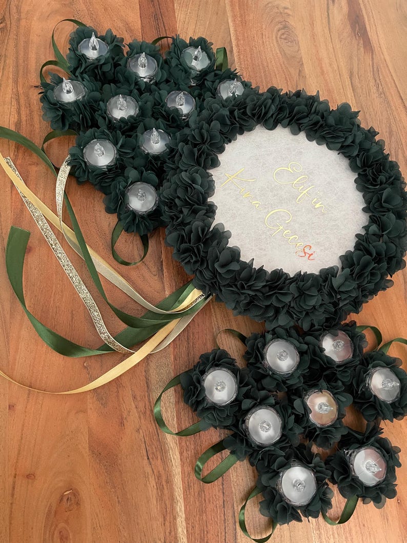 May include: Dark green floral arrangement with battery-operated lights and ribbons. The arrangement includes a central circular element with gold script. The design incorporates multiple clusters of dark green fabric flowers, each holding a small light.