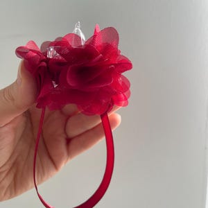 May include: A deep red floral wrist corsage with a sheer, layered petal design. The corsage features a clear, pointed embellishment at its centre and a matching red ribbon wrist strap.
