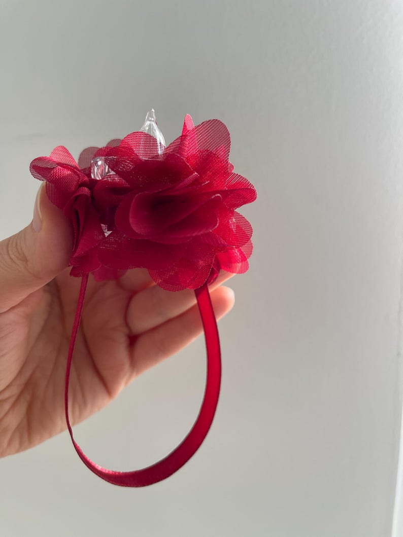May include: A deep red floral wrist corsage with a sheer, layered petal design. The corsage features a clear, pointed embellishment at its centre and a matching red ribbon wrist strap.