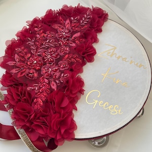 Könnte beinhalten: Eine runde, weiße dekorative Platte mit goldener Schrift "Zehra'nın Kına Gecesi". Ein Teil der Platte ist mit einer Kaskade aus tiefroten Stoffblumen, Perlen und Pailletten verziert. Ein gold-silbernes Band ist befestigt.