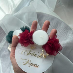 Op de afbeelding: Een witte ronde basis met de tekst "Selin in Kinasi" in gouden letters. De basis heeft twee rode bloemen van stof en een kleine witte theelichtkaars in het midden.