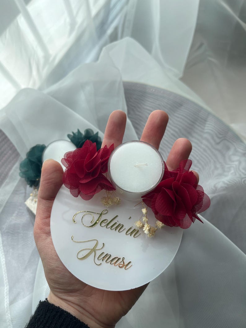 Op de afbeelding: Een witte ronde basis met de tekst "Selin in Kinasi" in gouden letters. De basis heeft twee rode bloemen van stof en een kleine witte theelichtkaars in het midden.
