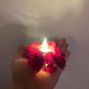 May include: A small LED candle with a flickering flame, encircled by deep red fabric flower petals. The candle is held in a hand, emitting a warm light. Ideal for decorative purposes.