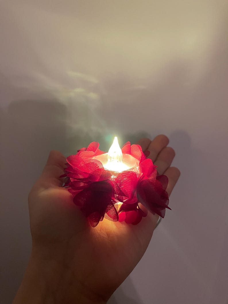 May include: A small LED candle with a flickering flame, encircled by deep red fabric flower petals. The candle is held in a hand, emitting a warm light. Ideal for decorative purposes.