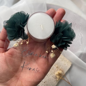 Op de afbeelding: Een ronde, transparante acrylplaat met de tekst "Esma'rin Kinasi" in zilveren letters. De plaat is versierd met twee groene stoffen bloemen en een witte theelichtje.