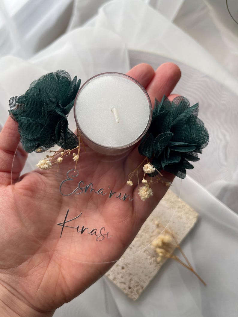Op de afbeelding: Een ronde, transparante acrylplaat met de tekst "Esma'rin Kinasi" in zilveren letters. De plaat is versierd met twee groene stoffen bloemen en een witte theelichtje.