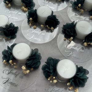 Op de afbeelding: Een set van heldere acryl kaarshouders met teal stoffen bloemen en witte theelichtjes. Elke houder heeft de tekst "Sedef in Kinası" erop geschreven.