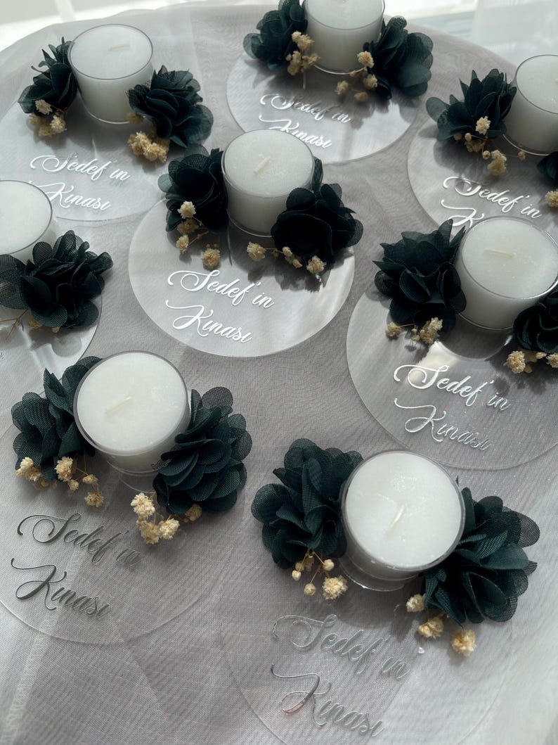 Op de afbeelding: Een set van heldere acryl kaarshouders met teal stoffen bloemen en witte theelichtjes. Elke houder heeft de tekst "Sedef in Kinası" erop geschreven.