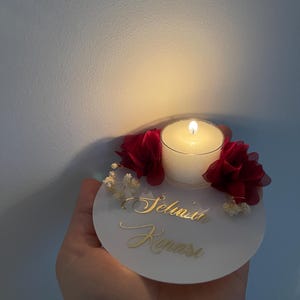 Op de afbeelding: Een ronde witte kaarsenhouder met een kleine witte kaars, een brandende lont en rode stoffen bloemen. De kaarsenhouder heeft de naam "Selina" in gouden letters.