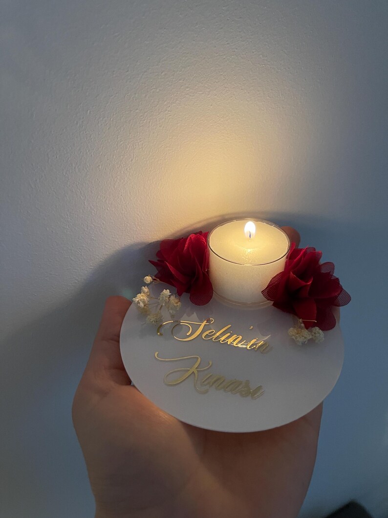 Op de afbeelding: Een ronde witte kaarsenhouder met een kleine witte kaars, een brandende lont en rode stoffen bloemen. De kaarsenhouder heeft de naam "Selina" in gouden letters.