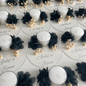 Op de afbeelding: Een set witte theelichthouders met zwarte bloemen van stof en witte gedroogde bloemen. De houders staan op een transparante acrylbasis met de tekst "Sedef in Kınası" erop.