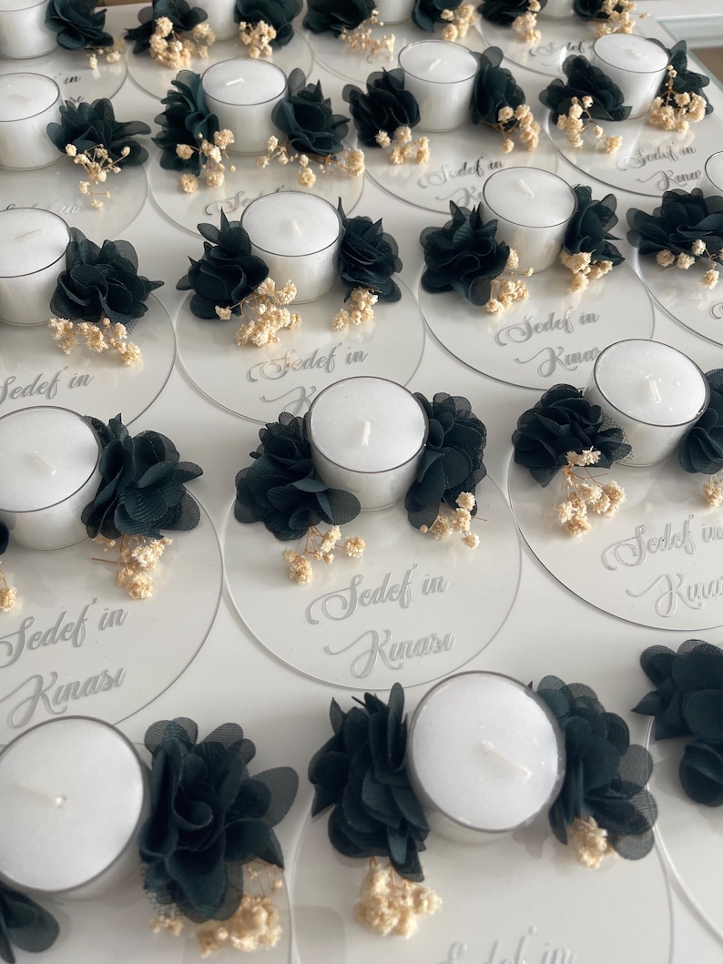 Op de afbeelding: Een set witte theelichthouders met zwarte bloemen van stof en witte gedroogde bloemen. De houders staan op een transparante acrylbasis met de tekst "Sedef in Kınası" erop.