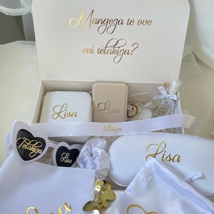 Könnte beinhalten: Eine weiße Geschenkbox mit einem weißen Schmuckkästchen, einem beigen Kompaktspiegel, einer kleinen Sprühflasche, herzförmigen Sonnenbrillen, einer weißen Schlafmaske und zwei weißen Beuteln. Alle Artikel tragen den goldenen Schriftzug Lisa oder Telakiga.