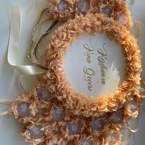May include: A peach-coloured floral wreath with a circular centre. The wreath is adorned with small, light-coloured flowers and decorative accents. The centre features gold script that reads "Kerban in Kina Gecesi". Ribbons and decorative elements are visible.