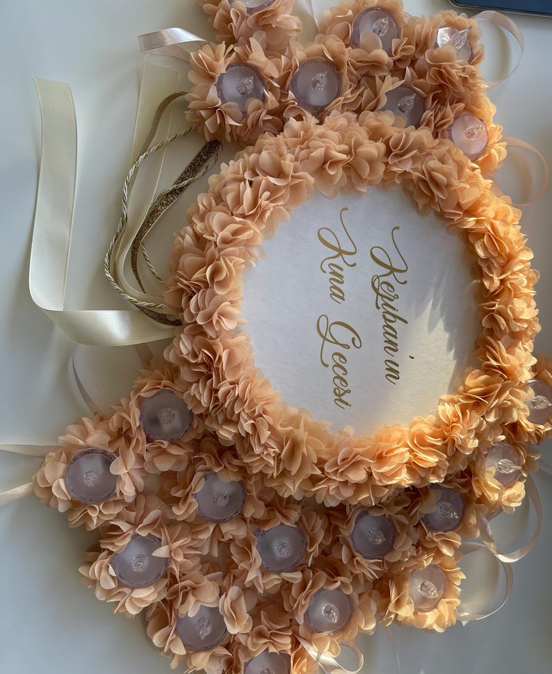 May include: A peach-coloured floral wreath with a circular centre. The wreath is adorned with small, light-coloured flowers and decorative accents. The centre features gold script that reads "Kerban in Kina Gecesi". Ribbons and decorative elements are visible.