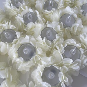 May include: Close-up of a floral arrangement featuring white fabric flowers surrounding clear, battery-operated LED tea lights. The arrangement is a soft white colour, with a delicate, layered petal design. Ribbons are visible.