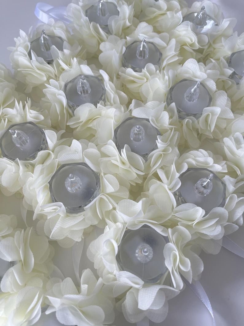 May include: Close-up of a floral arrangement featuring white fabric flowers surrounding clear, battery-operated LED tea lights. The arrangement is a soft white colour, with a delicate, layered petal design. Ribbons are visible.