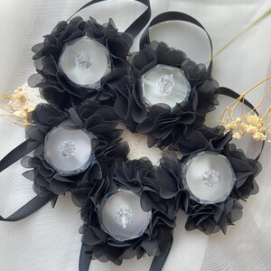May include: Five black floral hair accessories with clear, faceted centre stones. Each flower is made of layered, ruffled fabric and attached to a black ribbon. The accessories are arranged on a white, textured surface.