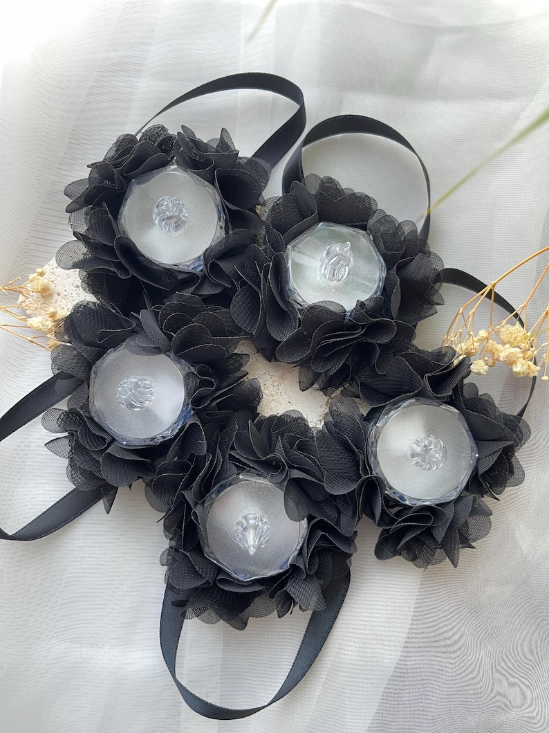 May include: Five black floral hair accessories with clear, faceted centre stones. Each flower is made of layered, ruffled fabric and attached to a black ribbon. The accessories are arranged on a white, textured surface.