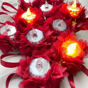 May include: A cluster of LED tea lights, each set within a red fabric flower. Some lights are lit, emitting a warm glow. Red ribbon adds to the decorative arrangement, creating a festive appearance.