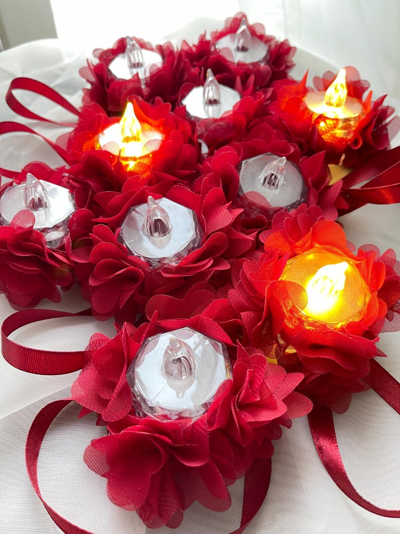 May include: A cluster of LED tea lights, each set within a red fabric flower. Some lights are lit, emitting a warm glow. Red ribbon adds to the decorative arrangement, creating a festive appearance.