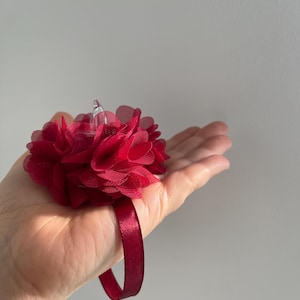 May include: A deep red wrist corsage with a cluster of layered fabric flowers. The floral arrangement is attached to a matching satin ribbon wristband. The corsage is burgundy in colour and is suitable for weddings, proms, or other special events.