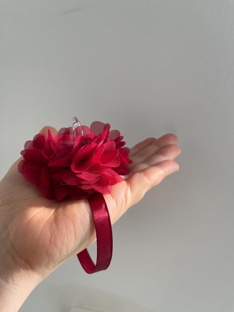 May include: A deep red wrist corsage with a cluster of layered fabric flowers. The floral arrangement is attached to a matching satin ribbon wristband. The corsage is burgundy in colour and is suitable for weddings, proms, or other special events.