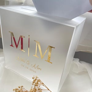 Könnte beinhalten: Eine rechteckige Kartenbox aus mattiertem Acryl mit einem Schlitz an der Oberseite. Die Vorderseite zeigt die goldfarbenen Buchstaben M|M, die Namen Melis & Mert in Schreibschrift und das Datum 03.10.2026. Ein weißer Umschlag wird in den Schlitz gesteckt.