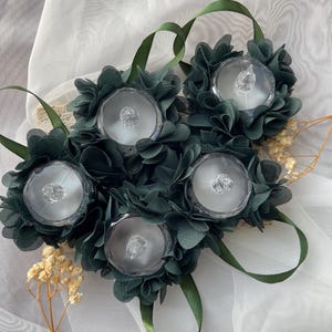 May include: Five dark green fabric flower wrist corsages. Each corsage has a clear, round, decorative centre, surrounded by dark green fabric petals. Green ribbon loops are attached for wearing. Dried flowers and white fabric are in the background.