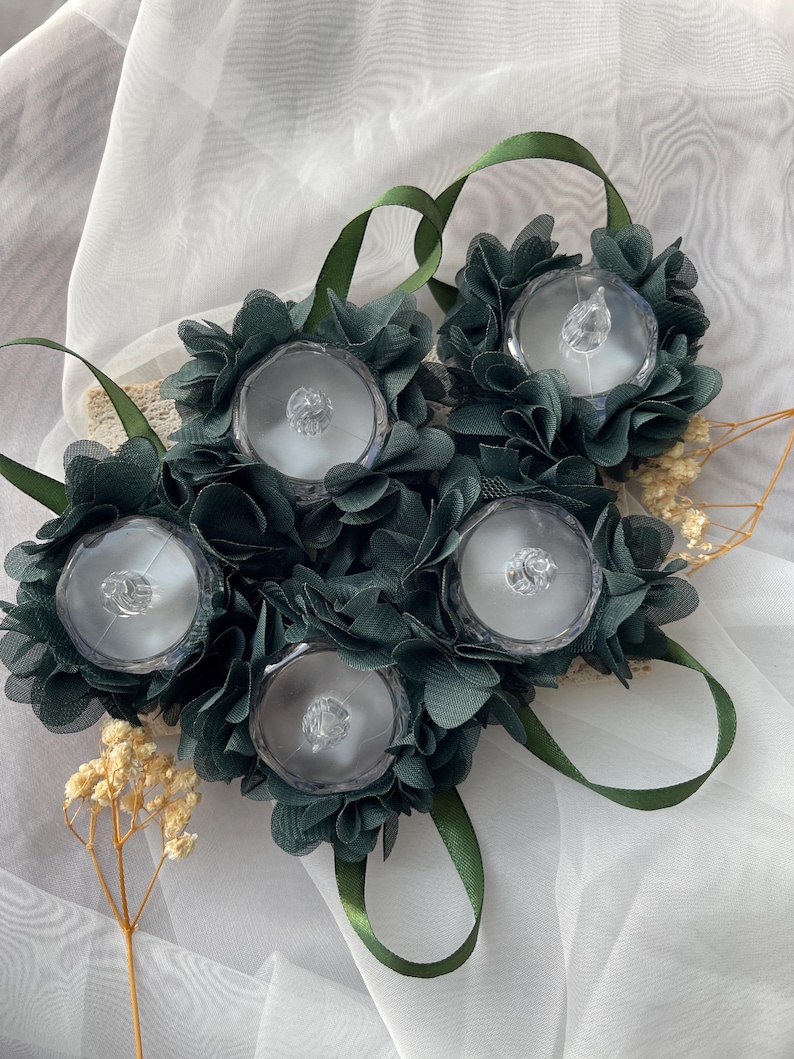 May include: Five dark green fabric flower wrist corsages. Each corsage has a clear, round, decorative centre, surrounded by dark green fabric petals. Green ribbon loops are attached for wearing. Dried flowers and white fabric are in the background.