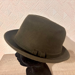 May include: A dark olive green fedora hat with a structured brim and a band detail. The hat is displayed on a black mannequin head. The hat's colour is consistent throughout, and it appears to be made of a felt-like material.